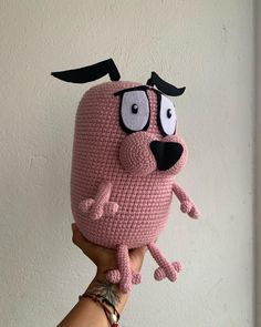 a hand holding up a crocheted pink object with black eyes and nose on it