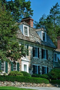 an old stone house with green shutters and trees in the front yard on a sunny day