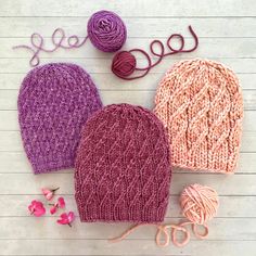 three knitted hats and yarn on a white wooden surface with pink flowers next to them