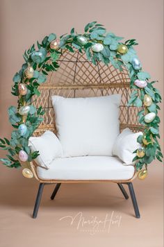 a wicker chair with white pillows and green leaves on it, sitting in front of a pink wall