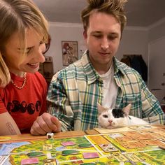 a man and woman looking at a board game with a cat sitting on the table