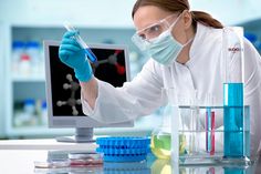 a woman in white lab coat and goggles holding up a test tube with blue liquid