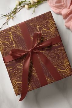 a gift wrapped in red and gold paper with a bow on it next to flowers