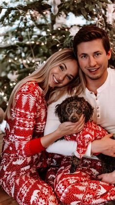 a man and woman sitting in front of a christmas tree with a dog on their lap