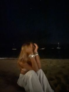 a woman sitting on the beach at night with her hands in her hair and looking off into the distance