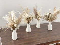 three white vases filled with dried flowers on top of a wooden table next to a wall