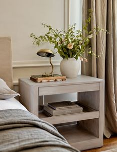 a white vase with flowers sitting on top of a small table next to a bed