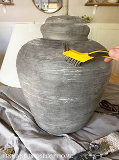 a person is holding a brush in front of a large gray vase on a table