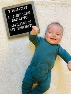 a baby laying on the floor holding up a sign that says 3 months i just like smiling, smiling is my favorite