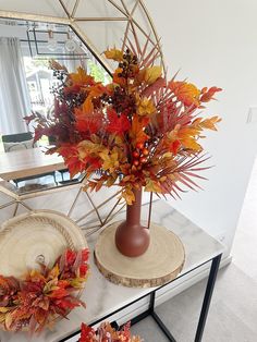 an arrangement of autumn leaves in a vase on a table with a mirror behind it