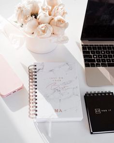 a desk with a laptop, notebook and flowers next to it on top of a white table