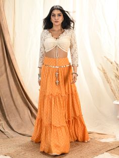 This is a three-piece mango bandhni lehenga set from the Aaryaa by KK collection. This beautiful silk mul tiered lehenga is paired with a floral lace blouse. The outfit is completed with a dupatta with coins tassel edging and it’s crafted in crush tissue material. The mango color with bandhni pattern, shells, and coin tassels enhance this lehenga set. The front tie-up style shimmer blouse has a V neckline with coin tassel embellishments and a back opening. Anarkali Choli With Dupatta And Tiered Skirt, Bollywood Style Choli With Dupatta And Tiered Skirt, Festive Bandhani Print Maxi Sets, Festive Bandhani Print Maxi Length Set, Designer Tiered Lehenga With Dupatta, Designer Anarkali Dress With Tiered Skirt, Festive Sharara With Dupatta And Tiered Skirt, Bohemian Style Fitted Lehenga For Eid, Bohemian Fitted Lehenga For Eid