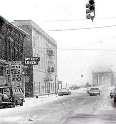 a black and white photo of cars driving down the street on a snow covered day