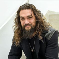 a man with long hair and beard wearing a black jacket sitting on a white bench