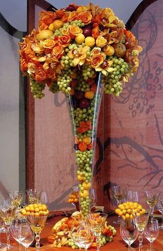 a tall vase filled with lots of fruit on top of a table covered in wine glasses