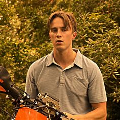 a young man standing next to an orange motorcycle in front of some bushes and trees