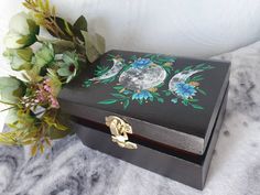 an ornate wooden box with painted flowers and moon on the lid sits next to a bouquet of greenery