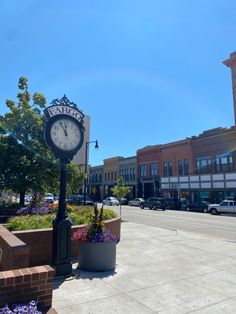 a clock on the side of a road with flowers in front of it and buildings behind it