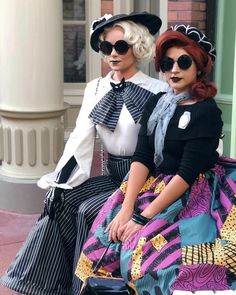 two women dressed in vintage clothing sitting on a bench with their hands behind their backs