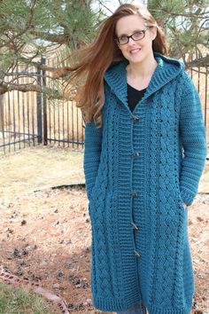 a woman standing in front of a fence wearing a blue knitted coat and glasses