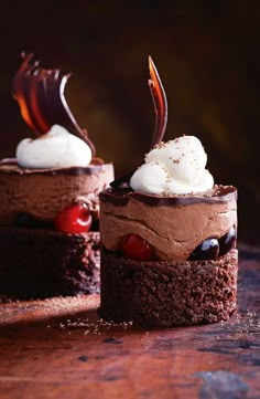 two chocolate cakes with whipped cream and cherries on top are sitting on a wooden table