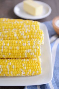 two corn cobs on a white plate next to butter and a blue striped table cloth