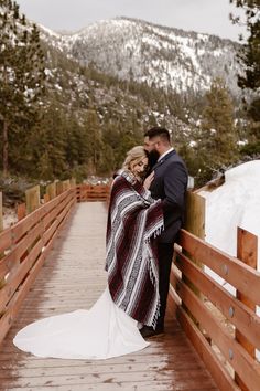 a bride and groom standing on a bridge in the snow with mountains in the background