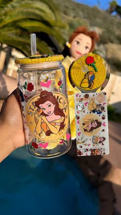 a hand holding up a glass jar with disney princess stickers on it and a doll next to it