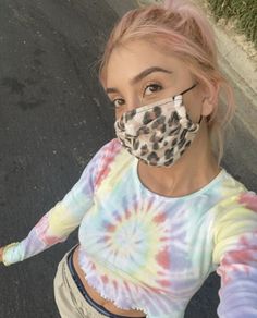 a woman with pink hair wearing a tie dye mask and looking at the camera while standing on a street