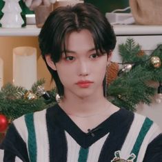 a young man with black hair wearing a green and white striped sweater standing in front of a christmas tree