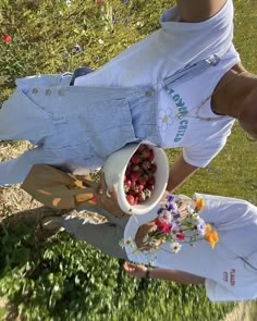 two women are picking strawberries out of a bowl on the grass in front of some flowers