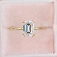 an emerald cut diamond sits in a pink velvet ring box, with the band around it's edge