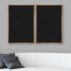 two black paintings hanging on the wall above a white couch in front of a gray wall