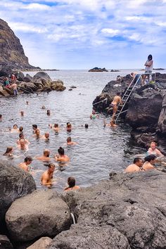 many people are swimming in the water near some large rocks and boulders, while one person stands on a ladder above them