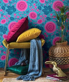 colorful pillows and blankets on a chair in front of a floral wallpaper