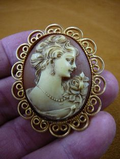 (Internal #CM43-53) Excellent, new, hand stained natural pine resin, hand-molded (reproduced from the original shell cameo) LADY WITH HAIR UP WITH ROSE FLOWER ON SHOULDER AND WEARING PEARLS AND EARRING CAMEO pin/pendant in IVORY color with BROWN background color, mounted on vintage filigree gold plated setting.  Cameo itself is 1-1/8" across x 1-1/2" long. Clasp pin/pendant glued on back. Superb detail. I know because I do half the work in creating the cameos. Comes with information card. WE SHI Elegant Carved Brooches For Gifts, Carved Brooches For Formal Occasions, Collectible Cabochon Brooches, Cameo Brooches For Wedding, Elegant Carved Gold Brooches, Ornate Carved Gold Brooches, Carved Gold Brooch For Gift, Ornate Gold Carved Brooches, Carved Gold Brooches As Gifts