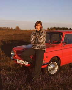 a woman sitting on the hood of a red car in a field with her legs crossed