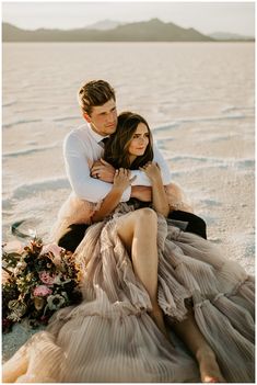 a man and woman are sitting on the sand in their wedding dresses, posing for a photo