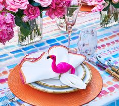 a flamingo napkin sits on an orange plate with pink flowers in vases behind it