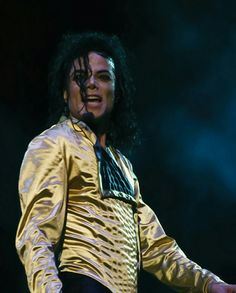 a man with long black hair wearing a gold jacket and bow tie, standing on stage