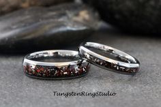 two wedding bands with red and black stones on them sitting next to each other in front of some rocks