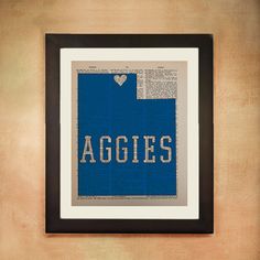 the word aggies written in blue and white is framed on a tan wall with a black frame