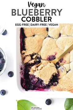 blueberry cobbler in a white dish with a gold serving spoon and green leaves