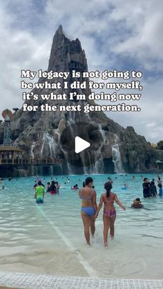 two women in bathing suits walking through the water at an amusement park with a waterfall behind them