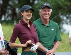 an older man standing next to a young woman on top of a green field with trees in the background