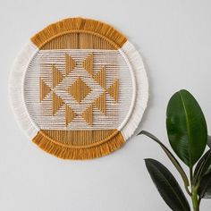 a white and yellow woven wall hanging next to a potted plant