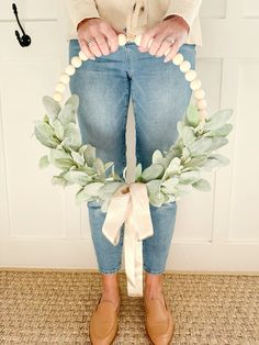 a woman is holding a wreath made out of wood beads and greenery with a white ribbon