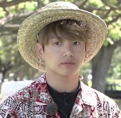 a young man wearing a straw hat standing in front of a park with trees and grass