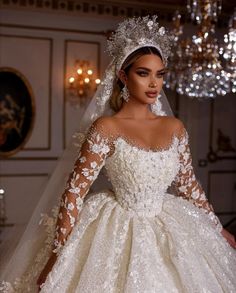 a woman wearing a wedding dress with long sleeves and flowers on the skirt is standing in front of a chandelier