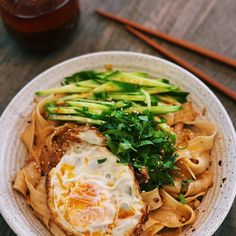 a white bowl filled with noodles and vegetables next to chopsticks on a wooden table